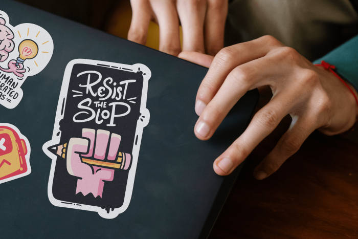 Close-up laptop mockup with a hand applying a hand-drawn “Resist the Slop” sticker featuring a raised fist holding a pencil. Surrounding stickers include human-generated ideas and anti AI creative badges.